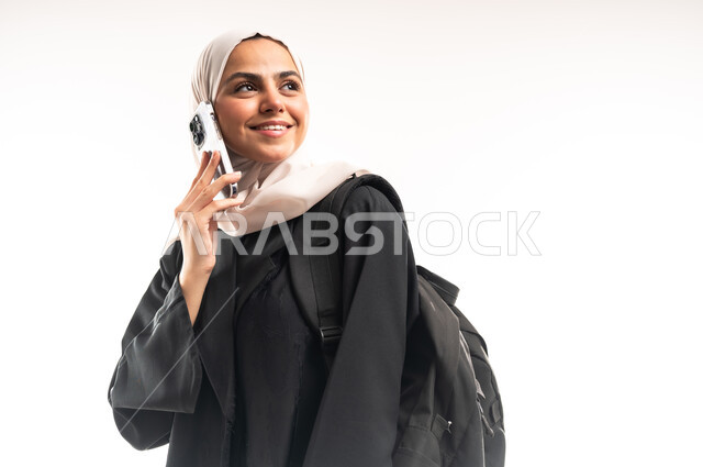 استخدام الأجهزة التقنية الحديثة في الحياة اليومية، إجراء اتصال هاتفي عبر الجوال, التعليم الجامعي في المملكة، بورتريه مقرب لطالبة جامعية عربية خليجية سعودية محجبة ترتدي عباءة سوداء تحمل حقيبة الظهر تمسك بيدها الهاتف المحمول، خلفية بيضاء