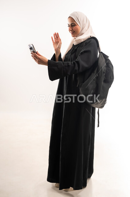 إجراء مكالمة فيديو عبر الجوال, استخدام الأجهزة التقنية الحديثة في الحياة اليومية، التعليم الجامعي في المملكة، بورتريه من الجانب لطالبة جامعية عربية خليجية سعودية محجبة ترتدي عباءة سوداء تحمل حقيبة الظهر تمسك بيدها الهاتف المحمول، صورة لكامل الجسم، خلفية بيضاء