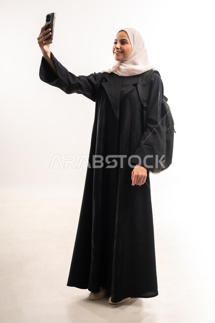 التقاط صور السيلفي التذكارية، استخدام الأجهزة التقنية الحديثة في الحياة اليومية، التعليم الجامعي في المملكة، بورتريه لطالبة جامعية عربية خليجية سعودية محجبة ترتدي عباءة سوداء تحمل حقيبة الظهر تمسك بيدها الهاتف المحمول، صورة لكامل الجسم، خلفية بيضاء