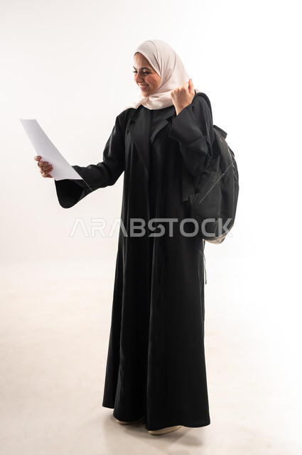 تحقيق الغايات والطموحات، الجامعات والكليات الأكاديمية، الاستعداد للعام الجامعي الجديد، بورتريه لطالبة جامعية عربية خليجية سعودية محجبة ترتدي عباءة سوداء تحمل حقيبة الظهر تمسك بورقة ترفع يدها بايماءات النصر والفوز، صورة لكامل الجسم، خلفية بيضاء