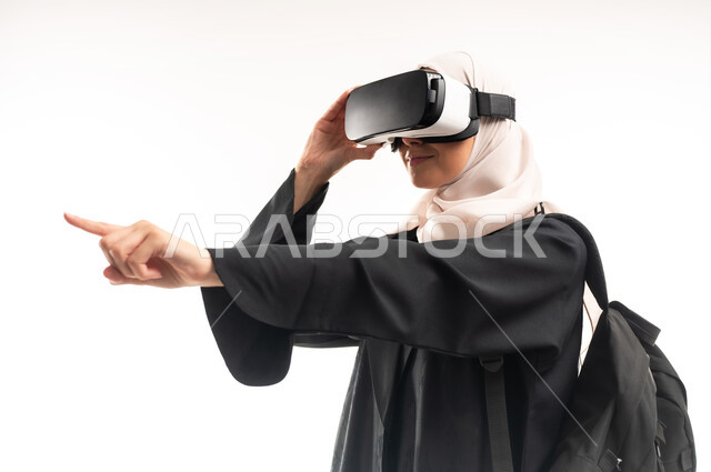 دمج التقنيات الحديثة بالتعلم، التعليم باستخدام تقنية العالم الافتراضي، مفهوم التفاعل والاندماج، بورتريه مقرب من الجانب لطالبة جامعية عربية خليجية سعودية محجبة ترتدي حقيبة الظهر تضع نظارة الفي ار ثلاثية الأبعاد تشير باصبع السبابة لمكان ما، خلفية بيضاء