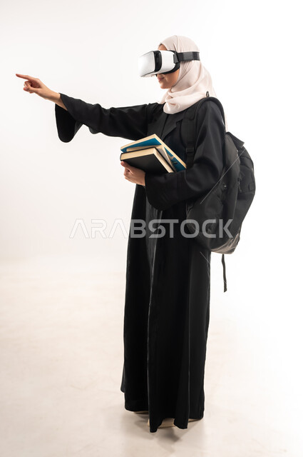 Concept of interaction and integration, simulation of the imaginary world, integration of modern technologies with learning, side portrait of a veiled Saudi Arabian Gulf university student wearing a backpack, 3D VR glasses, holding a set of books, pointing to a place with her hand, full body image, white background