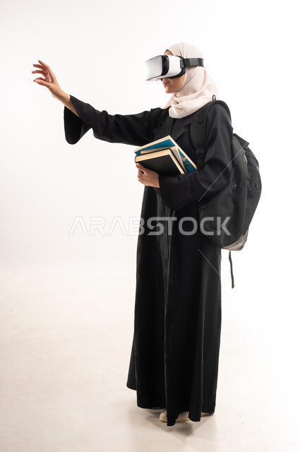 مفهوم التفاعل والاندماج، محاكاة العالم الخيالي، دمج التقنيات الحديثة بالتعلم، بورتريه من الجانب لطالبة جامعية عربية خليجية سعودية محجبة ترتدي حقيبة الظهر تضع نظارة الفي ار ثلاثية الأبعاد تحمل مجموعة من الكتب تشير بيدها لمكان ما، صورة لكامل الجسم، خلفية بيضاء