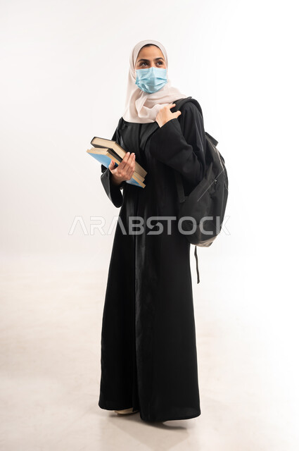 Modern curricula and educational materials, commitment to preventive measures, preparation for the new academic year, portrait of a veiled Saudi Arabian Gulf university student wearing a backpack and a medical mask, holding a group of books in her hand, full body image, white background