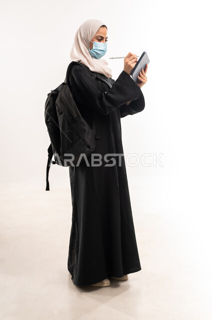 متابعة المحاضرات عبر المنصات التعليميه، مفهوم التعليم عن بعد، الالتزام بالاحترازات الوقائيه للحماية من فايروس كورونا، بورتريه من الجانب لطالبة جامعية عربية خليجية سعودية محجبة ترتدي الكمامة الطبية تحمل حقيبة الظهر وجهاز التابلت، صورة لكامل الجسم، خلفية بيضاء