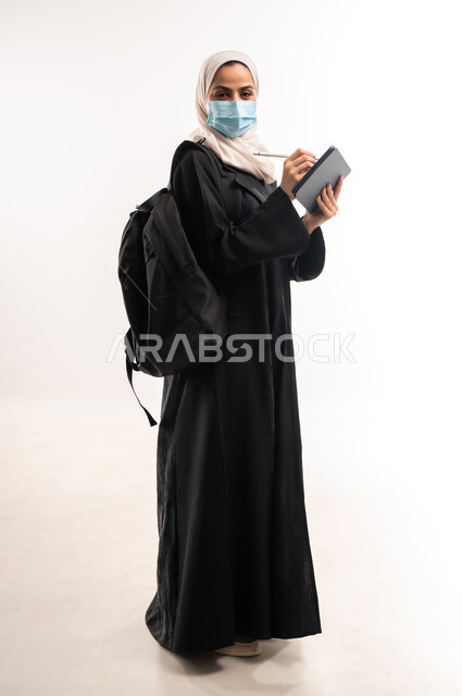 The concept of distance education, commitment to preventive precautions to protect against the Corona virus, following lectures via educational platforms, a side portrait of a veiled Saudi Arabian Gulf university student wearing a medical mask carrying a backpack and a tablet, a full body image, white background