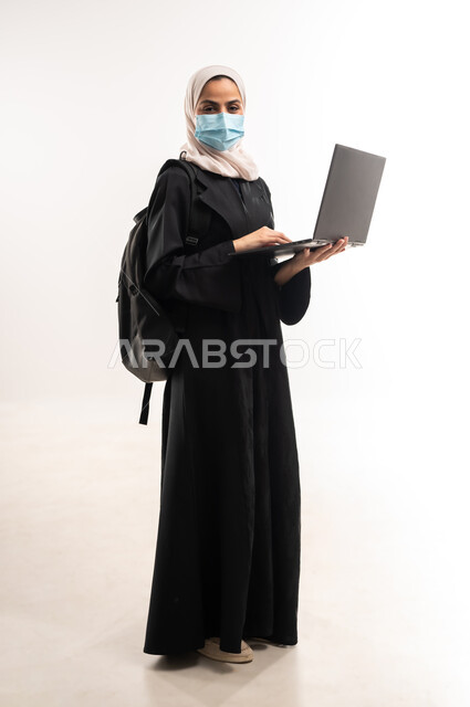 Using modern technical devices to follow lectures, the concept of distance education, a side portrait of a veiled Saudi Arabian Gulf university student wearing a medical mask carrying a backpack and a laptop, adhering to preventive precautions, a full body image, white background