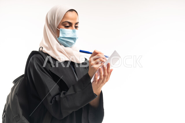 مفهوم اهمية التعليم في المملكة العربية السعودية، الالتزام بالاحترازات الوقائيه للحماية من فايروس كورونا، بورتريه مقرب من الجانب لطالبة جامعية عربية خليجية سعودية محجبة ترتدي الكمامة الطبية تحمل حقيبة الظهر تقوم بتسجيل وتدوين الملاحظات على الورق، خلفية بيضاء