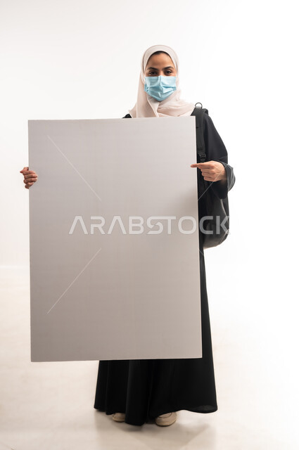 لوحات توعوية لتجنب إنتشار العدوى، الحفاظ على السلامة من كوفيد 19، اتباع الاجراءات الاحترازية للوقاية من فايروس كورونا، بورتريه لإمرأة عربية خليجية سعودية محجبة ترتدي الكمامة تنظر الى الكاميرا وتمسك بيدها موك اب مستطيل أبيض فارغ, الجسم الكامل بالطول، خلفية بيضاء