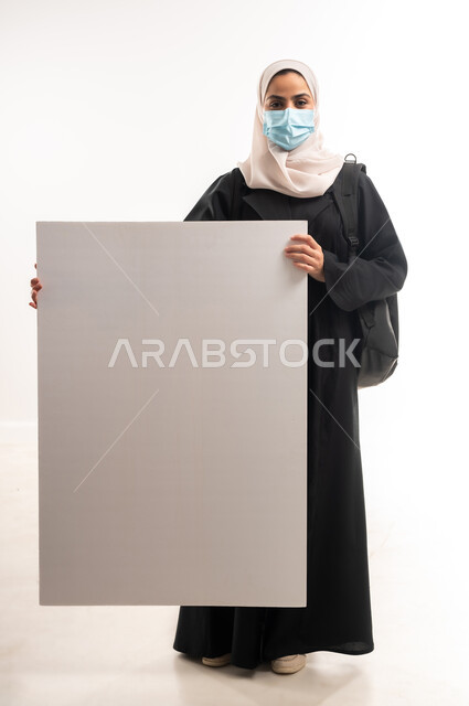 لوحات توعوية لتجنب إنتشار العدوى، الحفاظ على السلامة من كوفيد 19، اتباع الاجراءات الاحترازية للوقاية من فايروس كورونا، بورتريه لإمرأة عربية خليجية سعودية محجبة ترتدي الكمامة تنظر الى الكاميرا وتمسك بيدها موك اب مستطيل أبيض فارغ, الجسم الكامل بالطول، خلفية بيضاء