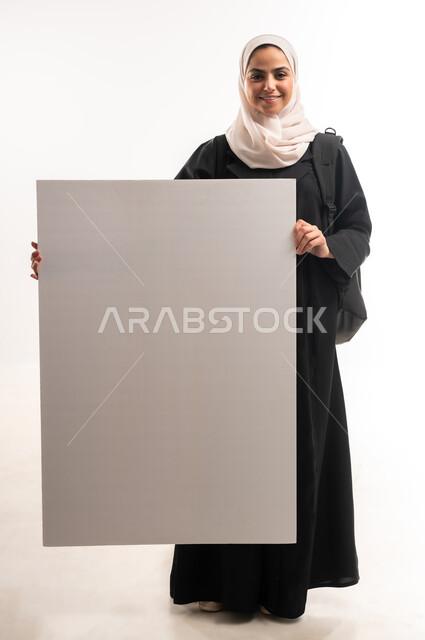 موك اب مستطيل ابيض، الاعلان عن بداية عام دراسي جديد في الجامعات السعودية الاكاديمية، بورتريه لطالبة جامعية عربية خليجية سعودية مبتسمة ترتدي الحجاب  حقيبة الظهر تمسك بيدها لوحة بيضاء فارغة بتعابير السرور، صورة الجسم بالكامل، خلفية بيضاء