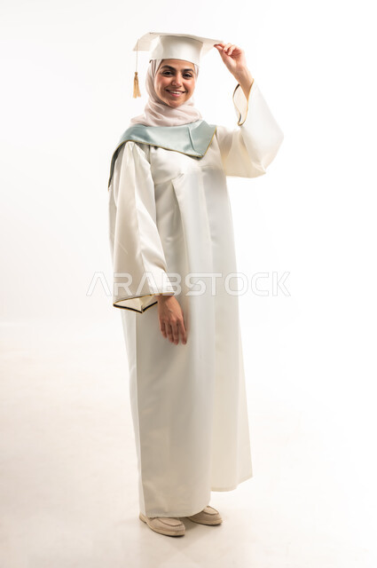 النظر الى الكاميرا بايماءات البهجة والسعادة بالوصول لحلم النجاح والتفوق، الاحتفال بإنهاء الدراسة الجامعية، التعليم في المملكة العربية السعودية, بورتريه لخريجة عربية خليجية سعودية محجبة مبتسمة ترتدي العباءة البيضاء والقبعة تمسك بيدها وثيقة التخرج، خلفية بيضاء