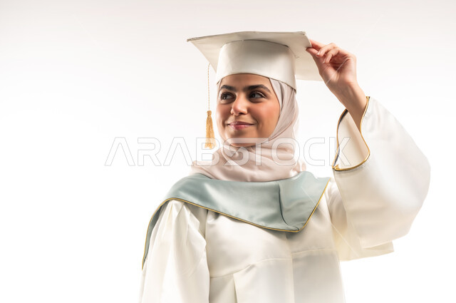 الاحتفال بإنهاء الدراسة الجامعية، التعليم في المملكة العربية السعودية, بورتريه مقرب لخريجة عربية خليجية سعودية محجبة مبتسمة ترتدي العباءة البيضاء وقبعة التخرج تنظر الى مكان ما بتعابير الفخر والثقة بالنفس، البهجة والسعادة بالوصول لحلم النجاح والتفوق، خلفية بيضاء