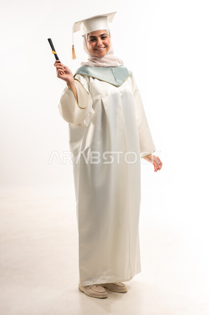 البهجة والسعادة بالوصول لحلم النجاح والتفوق، الاحتفال بإنهاء الدراسة الجامعية،, بورتريه لخريجة عربية خليجية سعودية محجبة مبتسمة ترتدي العباءة البيضاء والقبعة تحمل بيدها وثيقة التخرج، النظر الى الكاميرا بتعابير الفخر والسرور، الجسم الكامل بالطول،  خلفية بيضاء