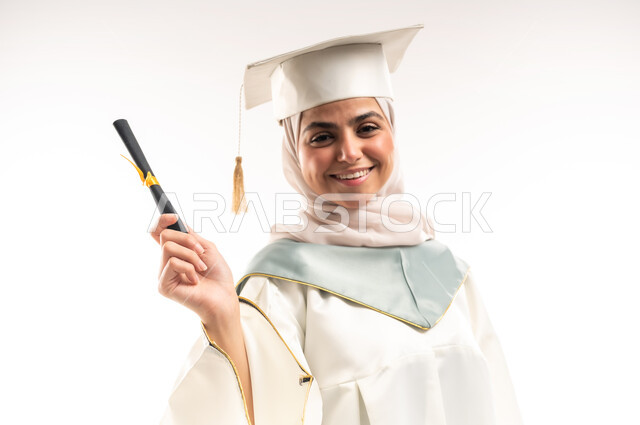 النظر الى الكاميرا بتعابير الفخر والسرور، السعادة بالوصول لحلم النجاح، الاحتفال بإنهاء الدراسة الجامعية، التعليم في المملكة العربية السعودية, بورتريه مقرب  لخريجة عربية خليجية سعودية محجبة مبتسمة ترتدي العباءة البيضاء والقبعة تحمل بيدها وثيقة التخرج، خلفية بيضاء