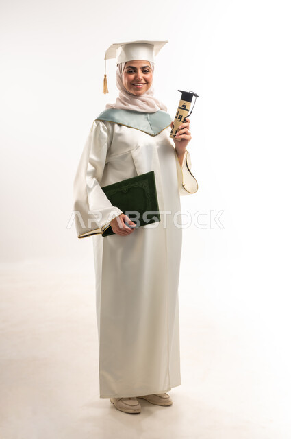 النظر الى الكاميرا بايماءات البهجة والسعادة بالوصول لحلم التخرج، الاحتفال بتحقيق النجاح، بورتريه لخريجة عربية خليجية سعودية محجبة مبتسمة ترتدي العباءة البيضاء والقبعة تمسك بيدها شهادة ووثيقة التفوق, صورة الجسم بالطول الكامل, خلفية بيضاء