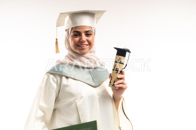 الاحتفال بإنهاء الدراسة الجامعية، التعليم في المملكة العربية السعودية, بورتريه مقرب لخريجة عربية خليجية سعودية محجبة مبتسمة ترتدي العباءة البيضاء والقبعة تمسك بيدها وثيقة التخرج، النظر الى الكاميرا بايماءات البهجة والسعادة بالوصول لحلم النجاح والتفوق، خلفية بيضاء