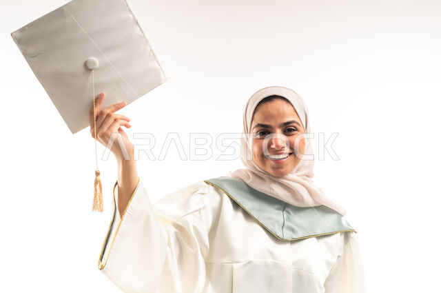 مفهوم النجاح والتفوق، الاحتفال بإنهاء الدراسة الجامعية، التعليم في المملكة العربية السعودية, بورتريه لخريجة عربية خليجية سعودية محجبة مبتسمة ترتدي العباءة البيضاء تمسك بيدها قبعة التخرج، النظر الى الكاميرا بايماءات البهجة والسعادة بالوصول لحلم انتهاء الدراسة، خلفية بيضاء