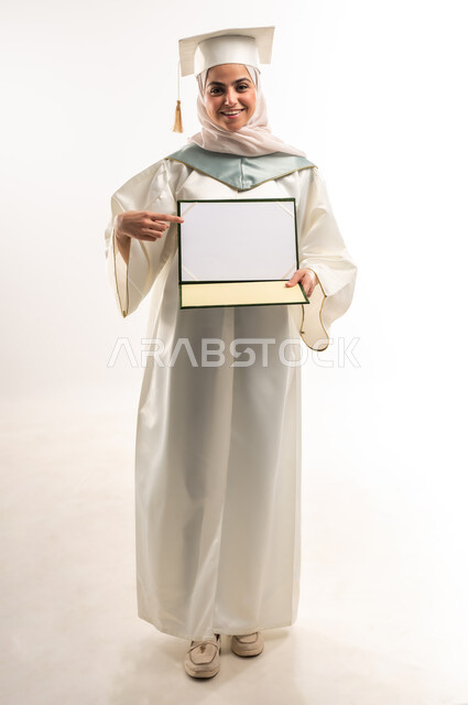الاحتفال بتحقيق النجاح, أهمية الدراسة الجامعية, ايماءات البهجة والسعادة بالوصول لحلم التخرج، بورتريه لخريجة عربية سعودية خليجية محجبة ترتدي العباءة البيضاء والقبعة تنظر الى الكاميرا تقوم بعرض شهادة التفوق, صورة الجسم بالطول الكامل, خلفية بيضاء