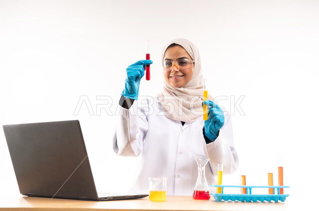 Integrating technology into practical life, close-up portrait of smiling Saudi Arabian Gulf female university student wearing white coat and glasses sitting behind her desk using test tubes to conduct scientific experiments in the laboratory, concept of biochemistry and research and development, white background