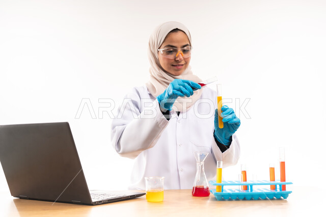 مفهوم الكيمياء الحيوية والبحث والتطوير، دمج الاجهزة الحديثة في الحياة العملية، بورتريه مقرب  لطالبة جامعية عربية خليجية سعودية ترتدي المعطف الابيض والنظارات الطبية تجلس خلف مكتبها تقوم باستخدام انابيب الاختبار لاجراء التجارب العلمية في المختبر،  خلفية بيضاء