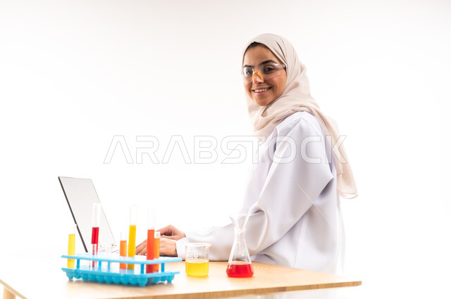 انابيب الاختبار لاجراء التجارب العلمية في المختبر، دمج الاجهزة الحديثة في الحياة العملية، بورتريه مقرب من الجانب لطالبة جامعية عربية خليجية سعودية ترتدي المعطف الابيض والنظارات الطبية  تقوم باستخدام جهاز اللابتوب،النظر الى الكاميرا بتعابي السرور، خلفية بيضاء