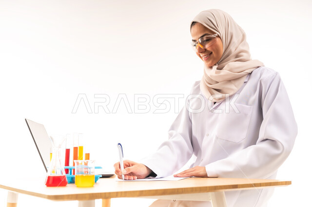 مفهوم الكيمياء الحيوية، بورتريه مقرب لطالبة جامعية عربية خليجية سعودية مبتسمة ترتدي المعطف الابيض والنظارات الطبية تقوم بتدوين وتسجيل نتائج التفاعلات الكيمائية على الاوراق في المختبر، دمج الاجهزة الحديثة في الحياة العملية، خلفية بيضاء