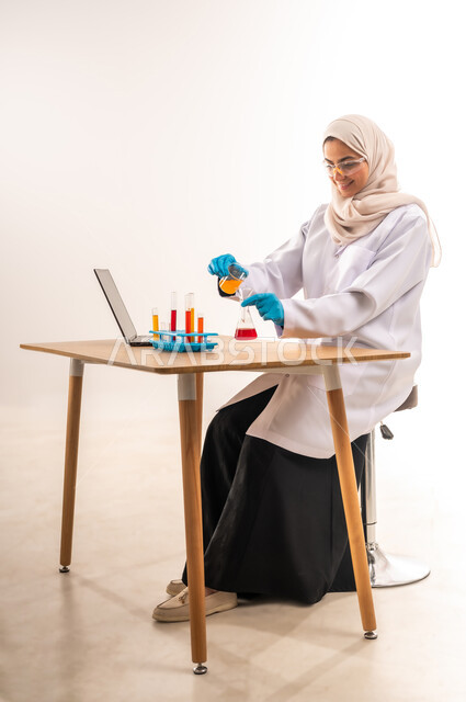 مفهوم الكيمياء الحيوية والبحث والتطوير، دمج الاجهزة الحديثة والتكنولوجيا في الحياة العملية، بورتريه لطالبة جامعية عربية خليجية سعودية مبتسمة ترتدي المعطف الابيض والنظارات الطبية  تقوم باستخدام انابيب الاختبار لاجراء التجارب العلمية في المختبر،  خلفية بيضاء