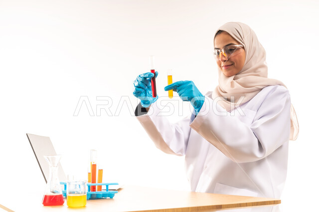 مفهوم الكيمياء الحيوية، البحث عن نتائج التفاعلات الكيميائية، استخدام الانابيب لاجراء التجارب العلمية، بورتريه مقرب لطالبة عربية خليجية سعودية مبتسمة ترتدي المعطف الابيض والنظارات الطبية تجلس في المخبر، دمج التعليم مع التقنية والتكنوجيا, خلفية بيضاء