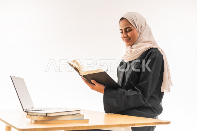 استخدام الأجهزة والتقنيات الحديثة والمتطورة, مفهوم التعليم عن بعد, بورتريه مقرب لطالبة جامعية عربية خليجية سعودية محجبة مبتسمة ترتدي العباءة تجلس على الطاولة وتقوم بتدوين المعلومات, مراجعة الدروس اليومية بواسطة اللابتوب، البحث الالكتروني عبر الانترنت, خلفية بيضاء