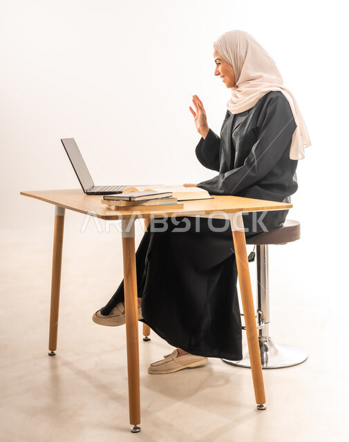 الاستعداد للمحاضرات الجامعية الالكترونية، مراجعة الدروس اليومية، استخدام الأجهزة التقنية الحديثة، ايماءات البشاشة والسرور، بورتريه لطالبة جامعية عربية خليجية سعودية محجبة ترتدي العباءة تجلس على الطاولة وتقوم بإجراء مكالمة فيديو عبر اللابتوب، خلفية بيضاء