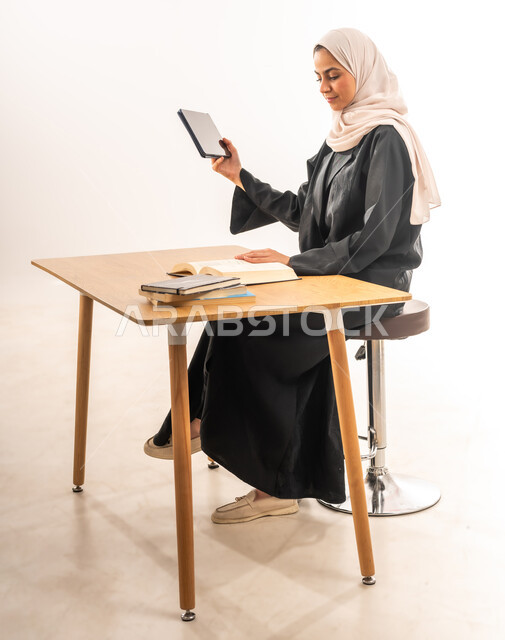 مراجعة الدروس اليومية بواسطة التابليت, البحث الالكتروني عبر الانترنت, استخدام الأجهزة والتقنيات الحديثة والمتطورة, مفهوم التعليم عن بعد, بورتريه لطالبة جامعية عربية خليجية سعودية محجبة مبتسمة ترتدي العباءة تجلس على الطاولة وتقوم بتدوين المعلومات, خلفية بيضاء