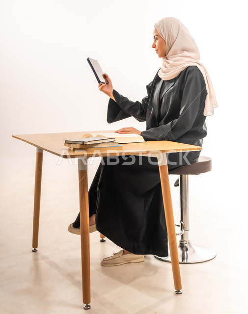 مفهوم التعليم عن بعد, بورتريه مقرب لطالبة جامعية عربية خليجية سعودية محجبة ترتدي العباءة تجلس على الطاولة وتقوم بتدوين المعلومات, مراجعة الدروس اليومية بواسطة التابليت، البحث الالكتروني عبر الانترنت, استخدام الأجهزة والتقنيات الحديثة والمتطورة, خلفية بيضاء