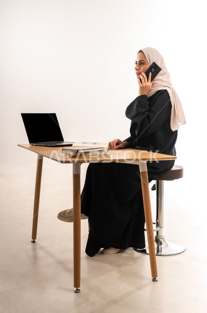 Chatting and communicating with friends, Portrait of a veiled, smiling, Saudi Gulf Arab university student wearing an abaya sitting at the table making a phone call via mobile phone, gestures of joy at the final exam results, using modern devices and technologies, white background