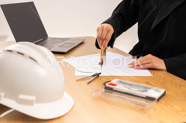 مفهوم الاجتهاد والتفوق, المناهج والمواد التعليمية, تصميم المشروع الهندسي، بورتريه مقرب لطالبة جامعية عربية خليجية سعودية محجبة ترتدي العباءة تجلس على الطاولة وتقوم بالرسم باستخدام الفرجار، دمج التقنيات الحديثة والتكنولوجيا بالتعليم الأكاديمي, خلفية بيضاء