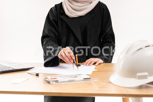 دمج التقنيات الحديثة والتكنولوجيا بالتعليم الأكاديمي, مفهوم الاجتهاد والتفوق, المناهج والمواد التعليمية, تصميم المشروع الهندسي، بورتريه مقرب لطالبة جامعية عربية خليجية سعودية محجبة ترتدي العباءة تجلس على الطاولة وتقوم بالرسم باستخدام الفرجار، خلفية بيضاء