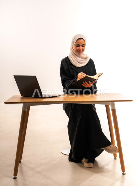 مراجعة الدروس اليومية, البحث الالكتروني عبر الانترنت, استخدام الأجهزة والتقنيات الحديثة والمتطورة, مفهوم التعليم عن بعد, بورتريه لطالبة جامعية عربية خليجية سعودية محجبة ترتدي العباءة تجلس على الطاولة وتقوم بتدوين المعلومات بواسطة جهاز اللابتوب, خلفية بيضاء