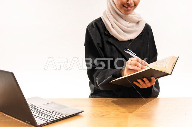 استخدام الأجهزة والتقنيات الحديثة, مفهوم التعليم عن بعد, بورتريه مقرب لطالبة جامعية عربية خليجية سعودية محجبة ترتدي العباءة تجلس على الطاولة وتقوم بتدوين المعلومات بواسطة جهاز اللابتوب, مراجعة الدروس اليومية, البحث الالكتروني عبر الانترنت, خلفية بيضاء