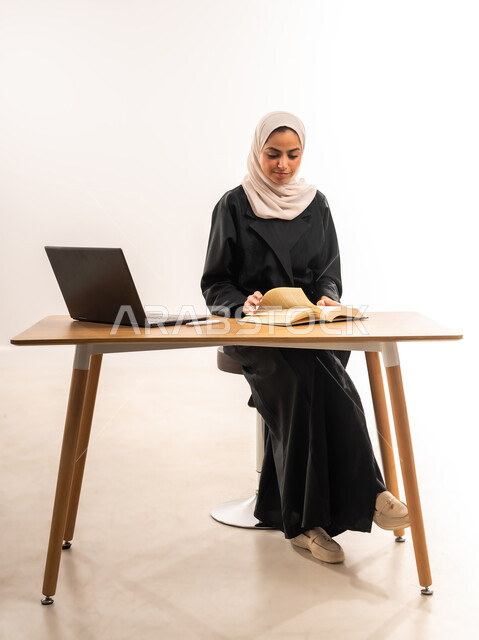 البحث الالكتروني عبر الانترنت, استخدام الأجهزة والتقنيات الحديثة والمتطورة, مفهوم التعليم عن بعد, بورتريه لطالبة جامعية عربية خليجية سعودية محجبة ترتدي العباءة تجلس على الطاولة وتقوم بتدوين المعلومات بواسطة جهاز اللابتوب, مراجعة الدروس اليومية, خلفية بيضاء