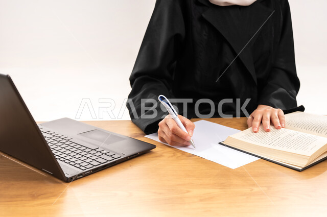 استخدام الأجهزة والتقنيات الحديثة, مفهوم التعليم عن بعد, بورتريه مقرب لطالبة جامعية عربية خليجية سعودية محجبة ترتدي العباءة تجلس على الطاولة وتقوم بتدوين المعلومات بواسطة جهاز اللابتوب, مراجعة الدروس اليومية, البحث الالكتروني عبر الانترنت, خلفية بيضاء