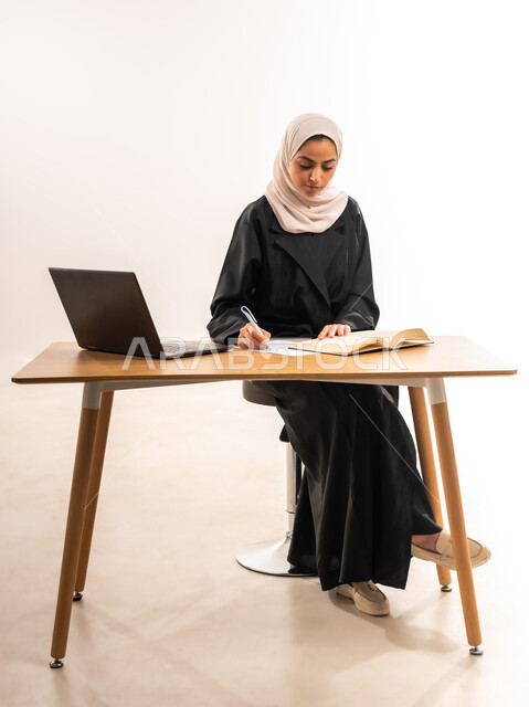 مراجعة الدروس اليومية, البحث الالكتروني عبر الانترنت, استخدام الأجهزة والتقنيات الحديثة والمتطورة, مفهوم التعليم عن بعد, بورتريه لطالبة جامعية عربية خليجية سعودية محجبة ترتدي العباءة تجلس على الطاولة وتقوم بتدوين المعلومات بواسطة جهاز اللابتوب, خلفية بيضاء