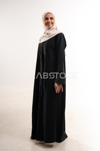 The concept of femininity and self-confidence, interest in elegance and appearance, portrait of a smiling veiled Saudi Arabian Gulf young woman wearing an abaya looking at something, gestures of happiness and pleasure, taking a selfie for official documents, standing straight and upright, full body length, white background