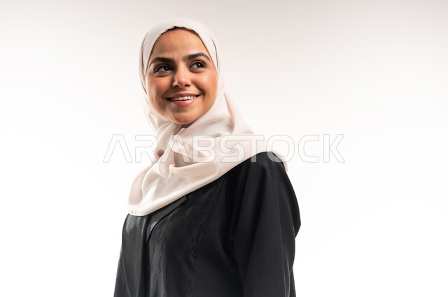 الوقوف باستقامة واعتدال، مفهوم الأنوثة والثقة بالنفس، الاهتمام بالأناقة والمظهر الخارجي، بورتريه مقرب لشابة عربية خليجية سعودية محجبة مبتسمة ترتدي العباءة السوداء وتنظر الى شيء ما، ايماءات السعادة والسرور، التقاط صورة شخصية للوثائق الرسمية، خلفية بيضاء