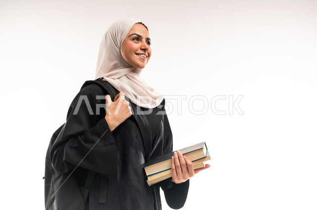 التعليم العالي في الجامعات والكليات الأكاديمية في السعودية, بورتريه مقرب لطالبة جامعية عربية خليجية سعودية محجبة ترتدي العباءة تضع حقيبة الظهر وتحمل الكتب, النظر الى شيء ما بإيماءات السعادة والسرور, الاستعداد للعام الجامعي الجديد, خلفية بيضاء