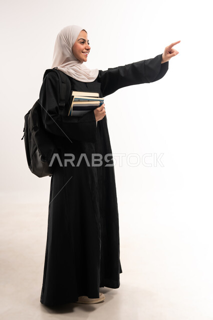 الإشارة بإصبع السبابة إلى شيء ما, الاستعداد للعام الجامعي الجديد بحماس, التعليم الاكاديمي في المملكة العربية السعودية, بورتريه لطالبة جامعية عربية خليجية سعودية محجبة ترتدي العباءة تضع حقيبة الظهر وتحمل الكتب, تحديد مكان معين، الجسم بالطول الكامل, خلفية بيضاء