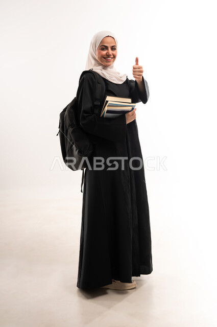 University education in Saudi Arabia, gestures of joy and admiration, culture and knowledge acquisition, portrait of a veiled Saudi Arabian Gulf university student wearing an abaya carrying a bag on her back holding books in her hand raising her thumb in a sign of satisfaction, full body length, white background