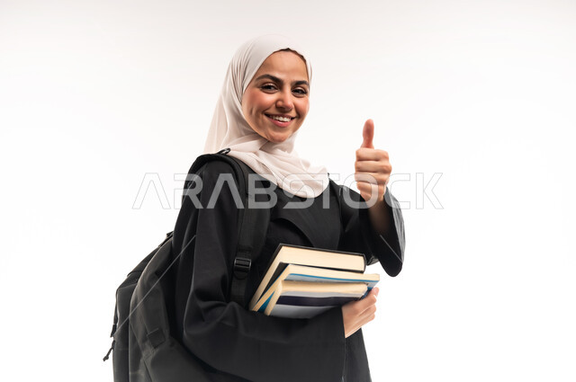 إيماءات الفرح والإعجاب لبداية العام الجديد، التثقف وإكتساب المعرفة، بورتريه مقرب لطالبة جامعية عربية خليجية سعودية محجبة ترتدي العباءة تضع حقيبة على ظهرها تحمل بيدها كتب ترفع اصبع الابهام بعلامة الرضا، التعليم الجامعي في المملكة العربية السعودية، خلفية بيضاء