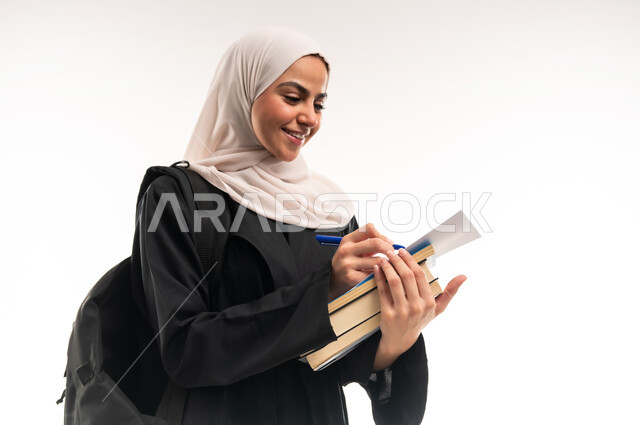 تدوين الملاحظات وحل الواجبات، مفهوم الاجتهاد والتفوق، بورتريه مقرب لطالبة جامعية عربية خليجية سعودية محجبة ترتدي العباءة تحمل حقيبة على ظهرها تحمل بيدها كتب، التثقف وإكتساب المعرفة، التعليم الجامعي في السعودية ، التحضير ومراجعة البحوث العلمية، خلفية بيضاء