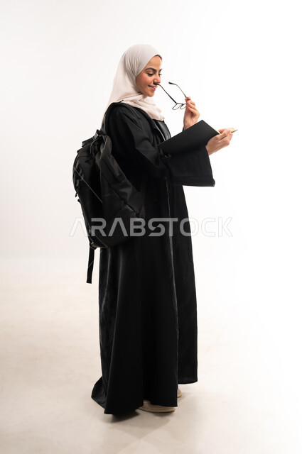 مراجعة الدروس اليومية, مفهوم الاجتهاد والتفوق, بورتريه من الجانب لطالبة جامعية عربية خليجية سعودية محجبة ترتدي العباءة تحمل حقيبة على ظهرها، تحضير البحوث العلمية, الاهتمام بتصفح الكتب المفيدة، التعليم الجامعي في المملكة, الطول الكامل للجسم، خلفية بيضاء