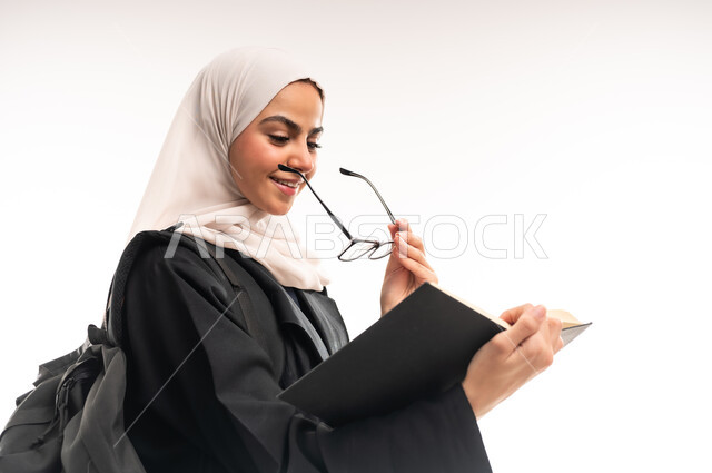 حب القراءة والمطالعة, بورتريه مقرب من الجانب لطالبة جامعية عربية خليجية سعودية محجبة ترتدي العباءة تحمل حقيبة على ظهرها وتضع النظارة الطبية، تحضير البحوث العلمية, الاهتمام بتصفح الكتب المفيدة، التعليم الجامعي في المملكة, مراجعة الدروس اليومية, خلفية بيضاء